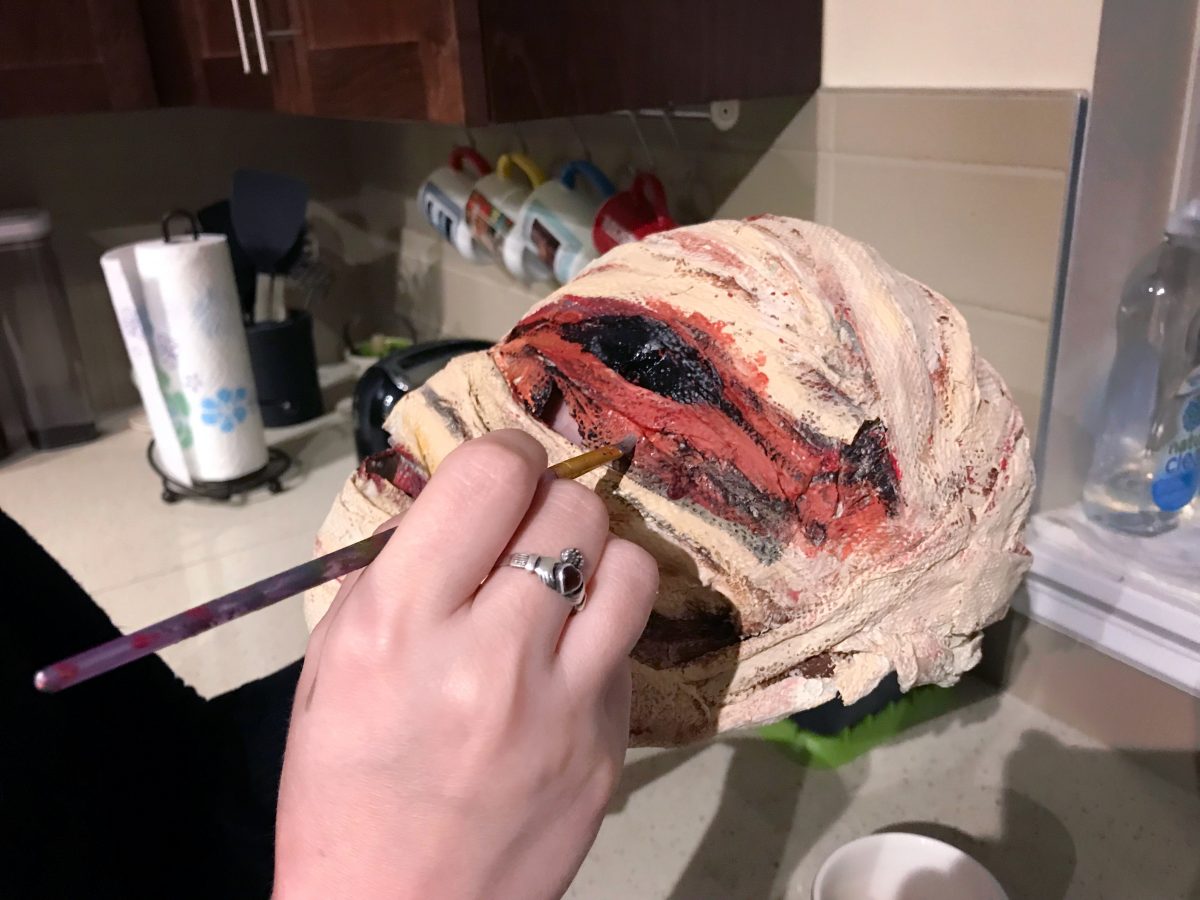 Tandye's hands in frame, holding a mask that appears to be nothing but bandages and blood. She is touching a paint brush to a bloody area under an empty eye hole.